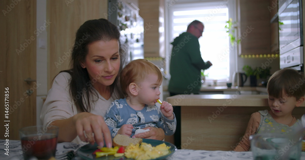 Obraz premium Mother interacting with children during breakfast, baby holding food while older sibling eats, warm family moment in cozy kitchen with everyday parenting connection
