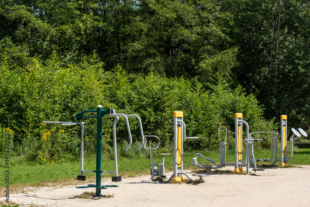 Fototapeta premium Empty outdoor fitness area with modern exercise machines in sunny green park surrounded by trees. Concept of healthy lifestyle, street workout, and public sport facilities in nature.
