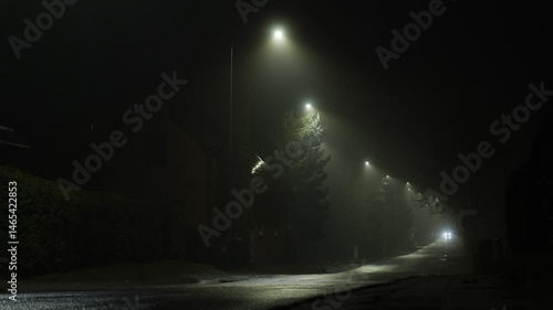 Moody foggy night street scene with a car passing under scattered streetlights.