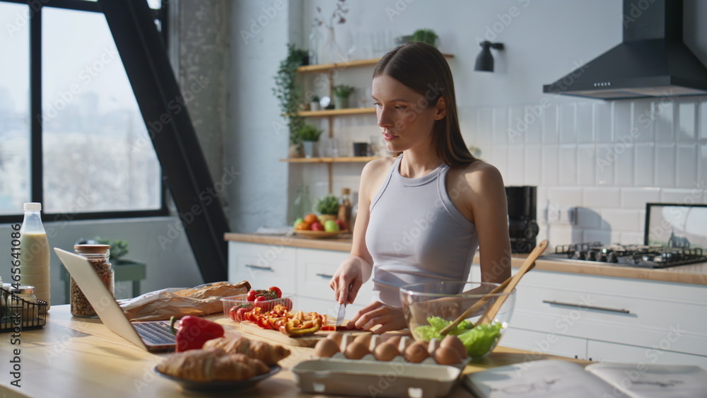 Serious woman chopping vegetables in modern kitchen watching serials on laptop