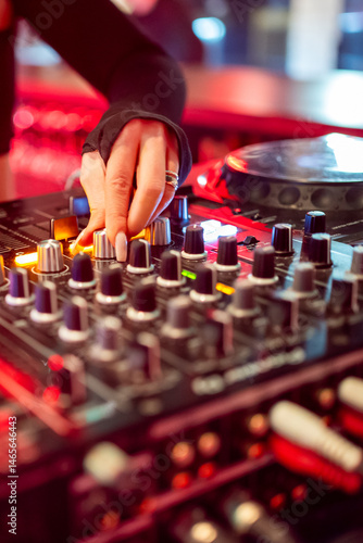 A close-up of a DJ's hand adjusting knobs on a professional audio mixer, illuminated by vibrant stage lighting. Captures the dynamic energy of a nightclub or live music performance.