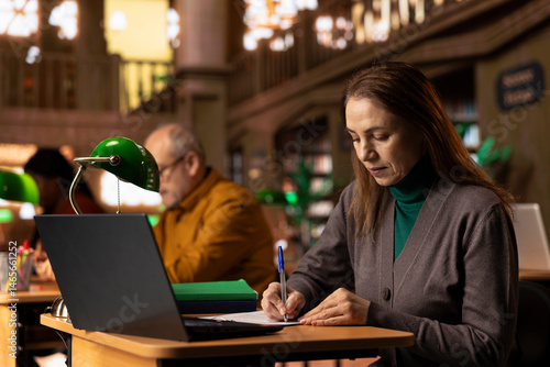 Obraz na płótnie Mature adult transcribing class work and reviewing materials for her university degree, surrounded by academic archived documents and books. Quiet atmosphere of learning in study area. w salonie