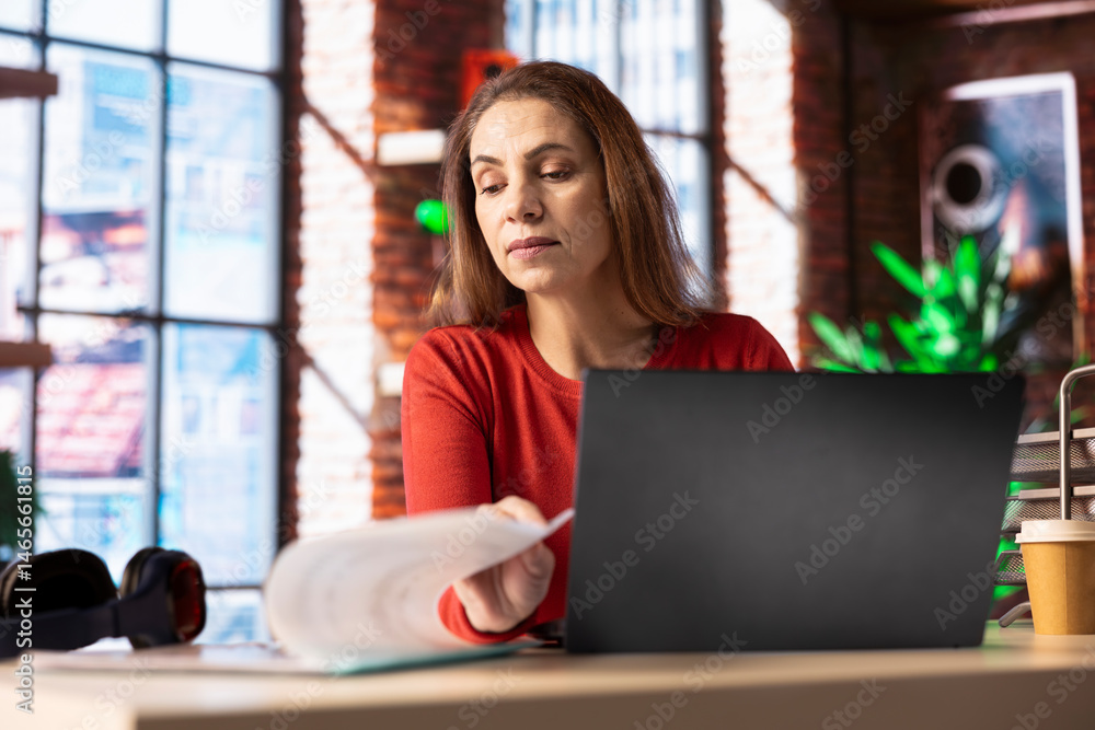 Casual person sitting at her desk and reading research papers, multitasking with laptop at home office. Woman diving into professional or personal topics of improvement, independent.