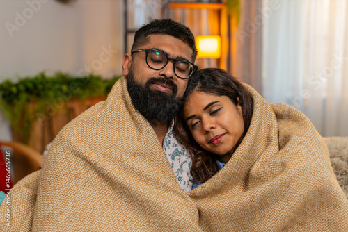 Indian couple man and woman sitting on home sofa, sharing soft hug under warm blanket in silence and comfort. Young family enjoying peaceful cozy moment together, feeling safe and emotionally close.