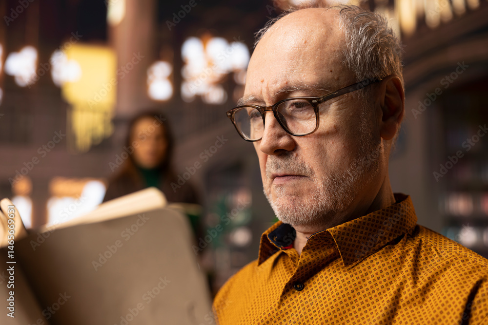 Retired man enjoys book reading at a public library and for leisure activity, feeling relaxed and increasing his knowledge during retirement. Happy calm person with eyeglasses, recreation.