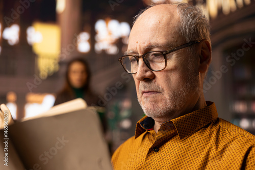 Obraz na płótnie Retired man enjoys book reading at a public library and for leisure activity, feeling relaxed and increasing his knowledge during retirement. Happy calm person with eyeglasses, recreation. w salonie