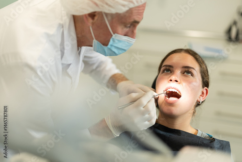Elderly male doctor dentist examining teeth of young female patient before treatment in office