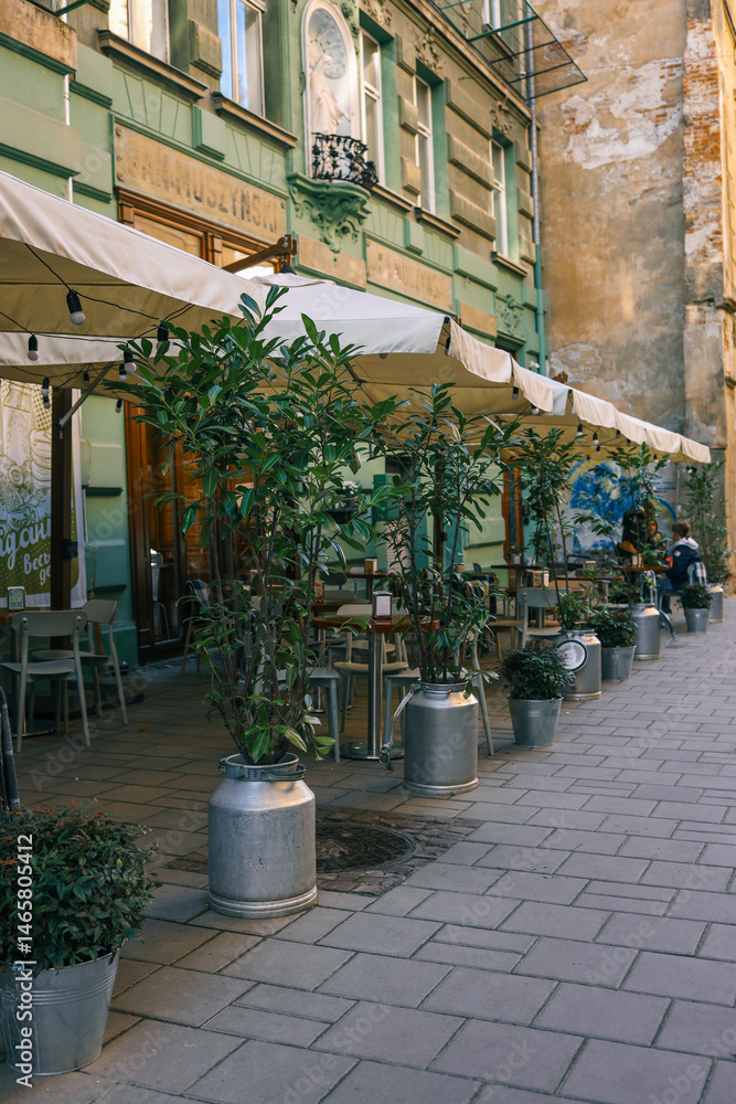 Obraz premium Outdoor café with cream awnings, metal pots with lush plants, and classic European architecture, creating a cozy and inviting street dining atmosphere