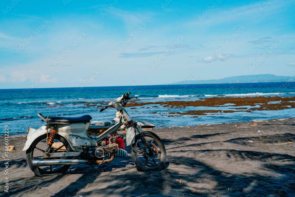 Fototapeta premium Vintage motorbike parked on a tropical beach with black sand and blue ocean in the background under clear sunny skies, evoking adventure and freedom