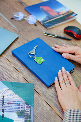 Close-up of a real estate workspace with a blue notebook, a house keychain, brochures, and a laptop on a wooden table. 