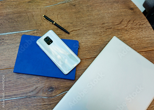 Top view of a wooden desk with a smartphone, a closed blue notebook, a pen, and a silver laptop. The modern phone and office supplies suggest a digital professional workspace. 