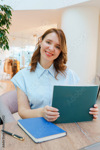 Portrait of a confident businesswoman sitting at a table in a modern office or coworking space. She is smiling warmly and looking at the camera while holding an open notebook. 