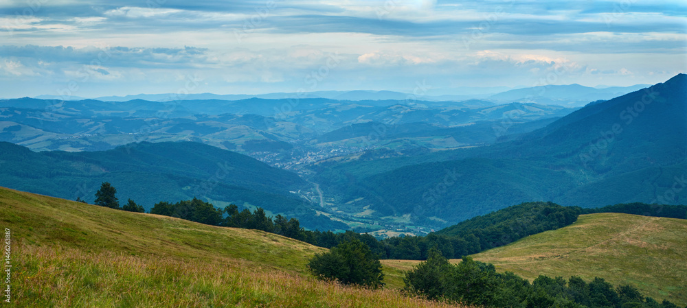 Fototapeta premium A sweeping panoramic view of verdant mountain ridges and valleys under a clear blue sky, with sunlit fields and distant villages nestled among lush green forests.