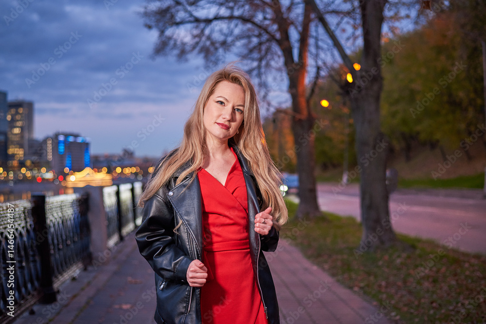 Fototapeta premium Woman in red dress is walking along embankment. She is wearing black jacket. Action takes place at night, with cityscape in background.