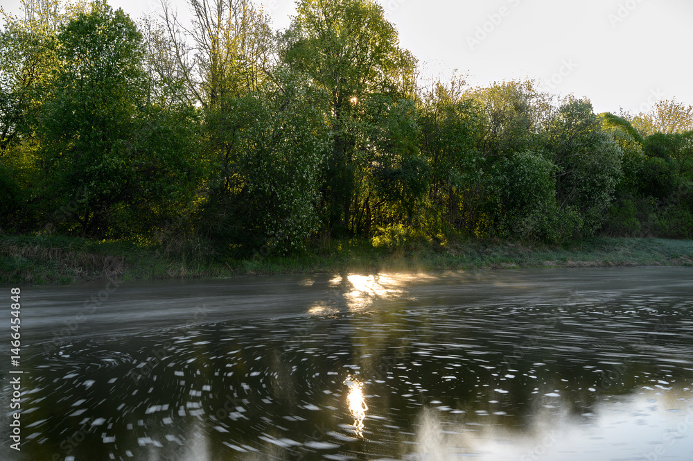Sunrise over a misty river surrounded by lush green trees in spring. Tranquil nature landscape with calm water, fog, and golden sunlight reflecting on the surface. Ideal for eco, travel, wellness.