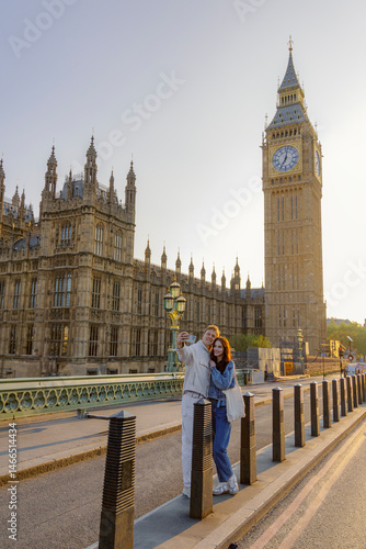A cheerful and happy couple is joyfully posing for a delightful selfie in front of the iconic Big Ben, capturing the essence of a beautiful, sunny day in the vibrant city of London