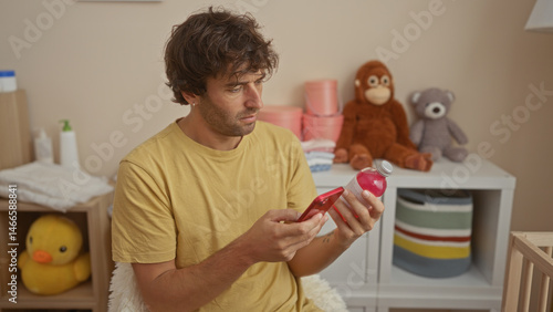 Young man in a bedroom holding a phone and a bottle, surrounded by baby toys, including a duck and stuffed animals, looking focused and attentive in a warm indoor setting.