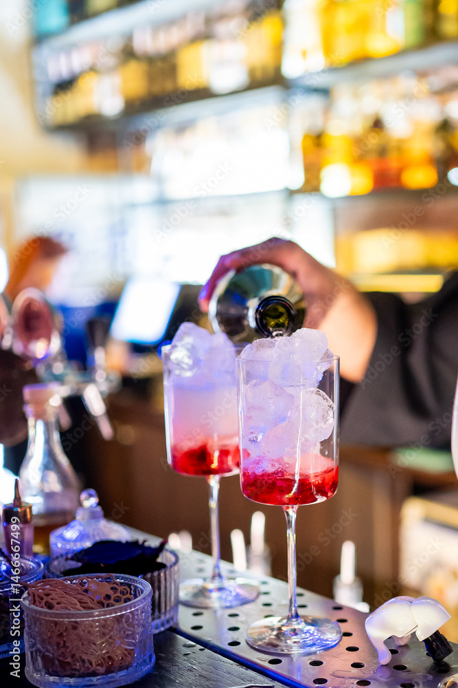 A bartender pours a cocktail into two tall glasses filled with ice and red syrup. The bar counter has various containers and tools, with blurred shelves of bottles in the background.
