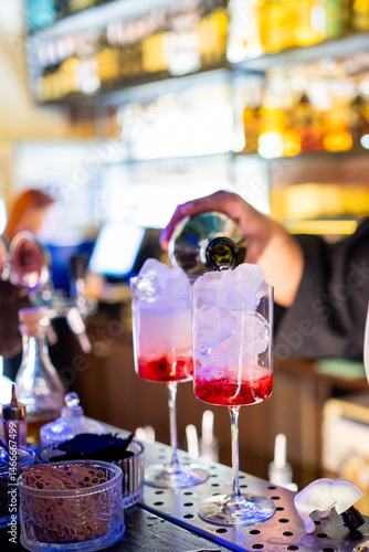 Obraz na płótnie A bartender pours a cocktail into two tall glasses filled with ice and red syrup. The bar counter has various containers and tools, with blurred shelves of bottles in the background. w salonie