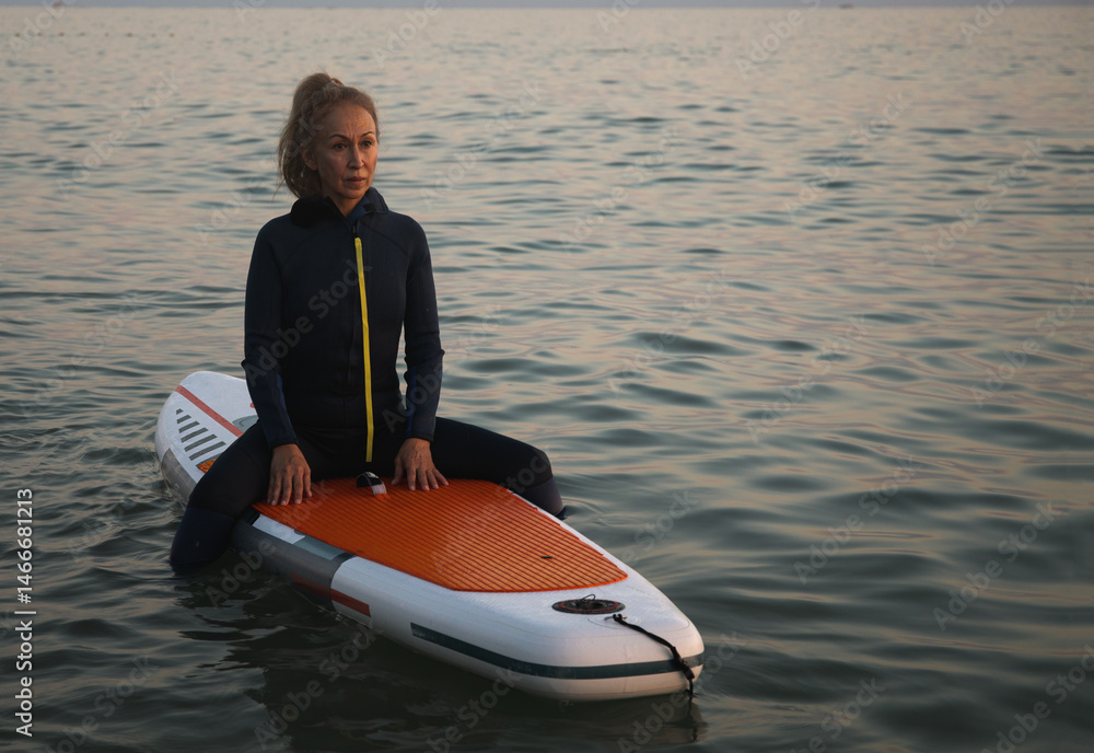 Obraz premium An elderly woman rides paddleboarding in the ocean. Senior female is waiting for a wave on a surfboard. Active old age. Old girl is sitting on a board in the sea at sunset, dressed in a swimming suit