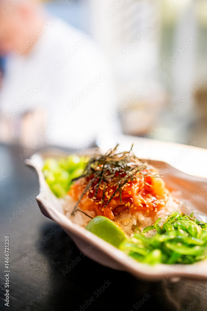 A vibrant poke bowl with fresh vegetables, rice, and seaweed, garnished with sesame seeds and lime, served on a dark surface. The colorful dish highlights textures and flavors in a gourmet presentatio