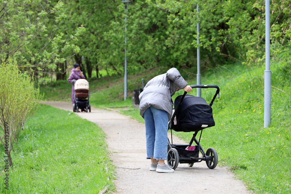 Mothers walking with baby prams in city park at spring. Woman with stroller in foreground