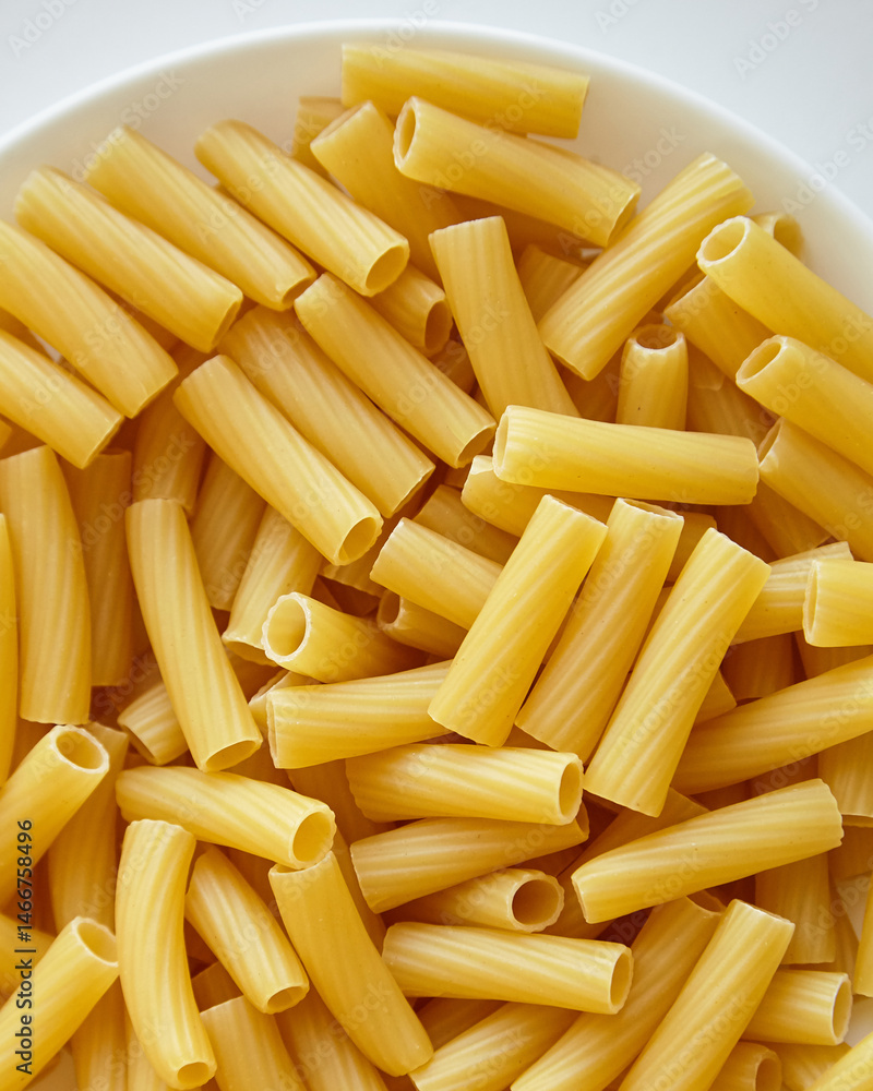 Overhead view of dry tortiglioni pasta in a round white bowl, showcasing its tubular shape and ribbed texture in natural lighting Overhead view of dry tortiglioni pasta in a round white bowl, showcasing its tubular shape and ribbed texture in natural lighting