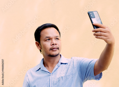 A man wearing a light blue shirt is standing and taking a selfie with a mobile phone. He raises his arm slightly forward to hold the phone and looks at the screen with a calm expression. 