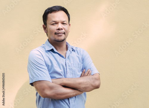 Asian man with a muscular build, wearing a light blue shirt, standing with arms crossed, looking at the camera with a confident expression; background is a plain light-colored wall.