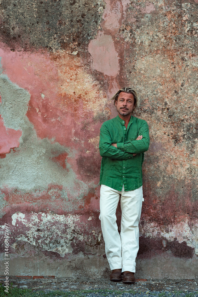 Obraz premium Tanned man with blonde hair in green shirt and white pants leaning against old pink stucco wall.