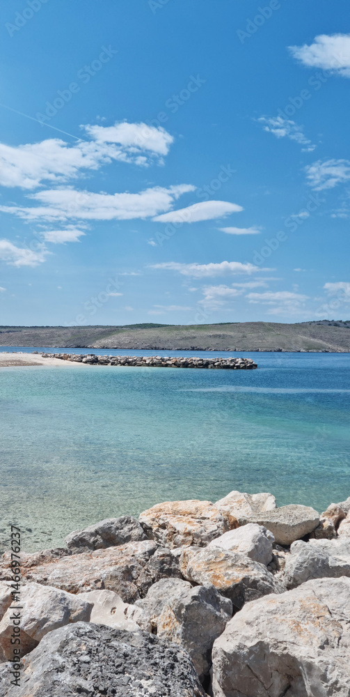 A scenic view from the rocky shore of Jadranovo overlooks the clear turquoise sea and the distant coastline of Krk island under a bright blue sky with scattered clouds