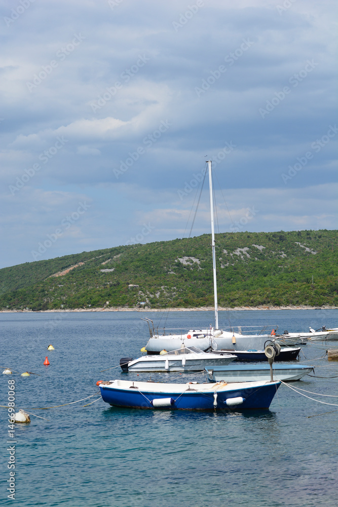 Several small boats, including a sailboat, are moored in the calm blue waters of Jadranovo near a green, hilly coastline under a partly cloudy sky