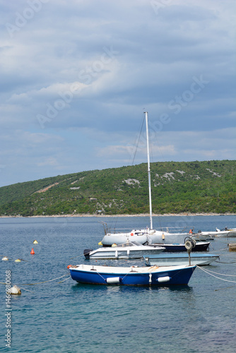 Obraz na płótnie Several small boats, including a sailboat, are moored in the calm blue waters of Jadranovo near a green, hilly coastline under a partly cloudy sky w salonie