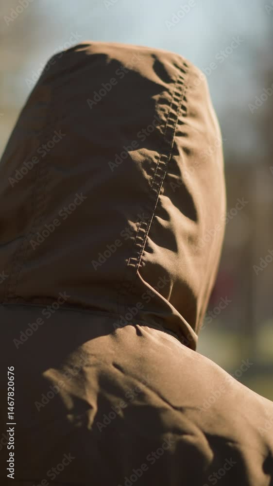A close- up of an individual wearing a brown hooded jacket walking outdoors, with a blurred background of trees and a pathway A close- up of an individual wearing a brown hooded jacket walking outdoors, with a blurred background of trees and a pathway
