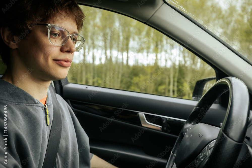 A young man with glasses and a grey hoodie smiles while driving a car along a road surrounded by trees. A young man with glasses and a grey hoodie smiles while driving a car along a road surrounded by trees.