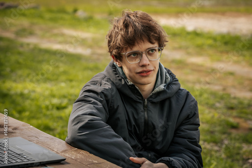 A young man in a black jacket and glasses sits by a wooden table outdoors, appearing thoughtful and relaxed in a grassy natural setting.