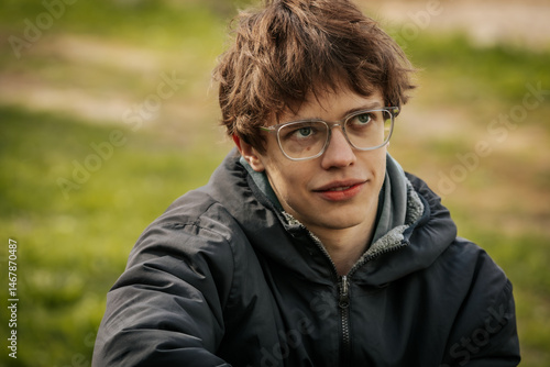 A young man in a black jacket and glasses sits by a wooden table outdoors, appearing thoughtful and relaxed in a grassy natural setting.