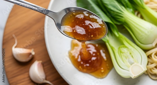 Pouring sauce from spoon onto plate with bok choy, noodles, and garlic cloves nearby