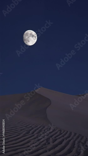 Full moon shining above silent desert dunes under deep blue night sky

