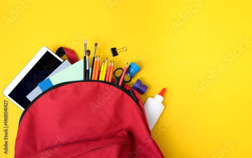 Red school backpack filled with glue, pencils, tablet, brushes, clips and notebooks on yellow. Great image for back to school sales, children’s education, and classroom promos.
