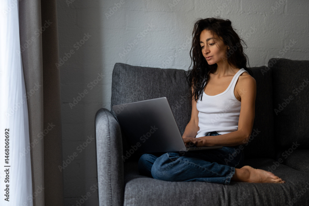 Naklejka premium Concentrated young woman sitting on a sofa, working on a laptop with natural light coming from the window in a modern minimalist room.