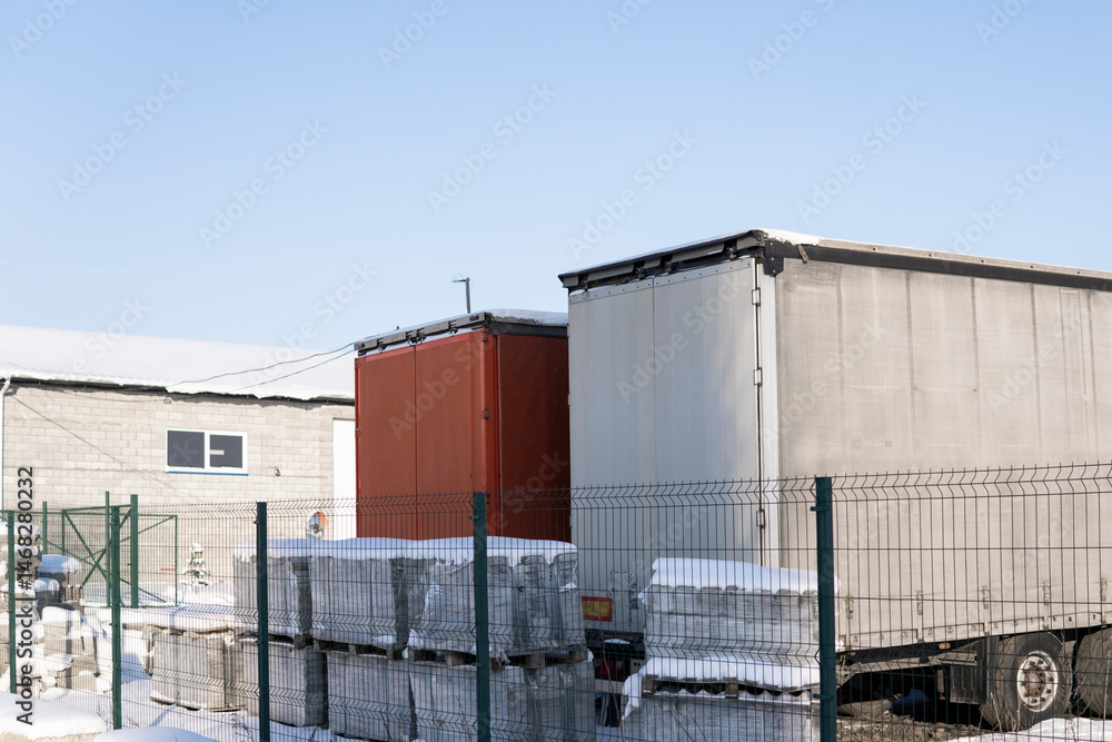 Obraz premium Snowy logistics yard with trailers, stacked pallets and industrial warehouse in sunlight