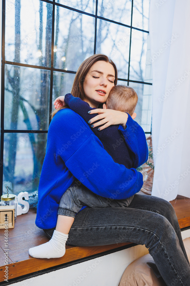 Happy mother and her 4 year old son hugging while sitting on the windowsill at home. Trusting relationship between mother and son. Little boy hugging his mother.