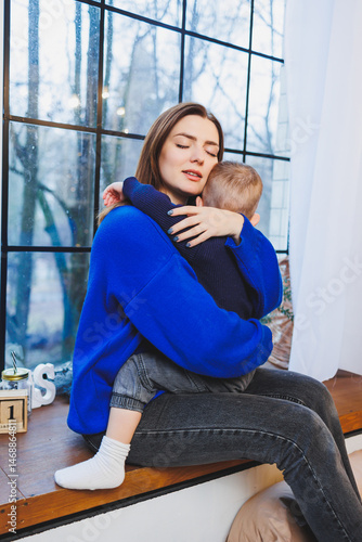 Obraz na płótnie Happy mother and her 4 year old son hugging while sitting on the windowsill at home. Trusting relationship between mother and son. Little boy hugging his mother. w salonie