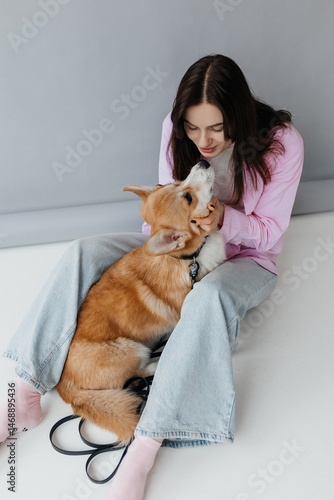 Obraz na płótnie Woman in jeans sitting on the floor and playing with a corgi dog. Cute brunette woman in a white t-shirt and jeans hugging a corgi on a white background. Love for animals. woman holding a corgi dog. w salonie