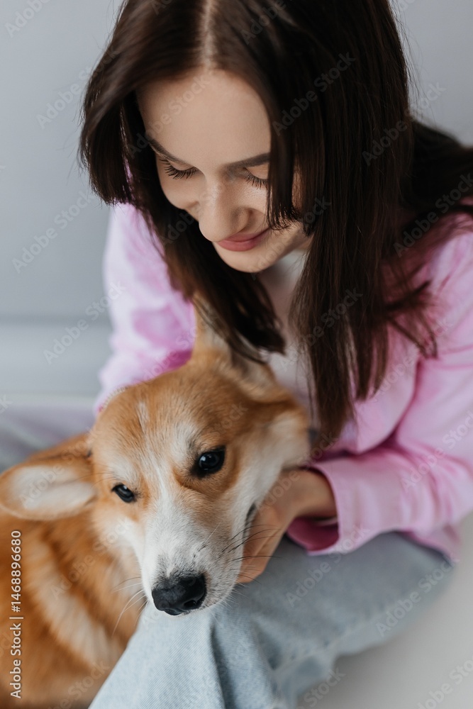 Happy brunette sitting next to corgi dog indoors. Cheerful girl in good mood stroking animal. Concept of friendship between man and pet.