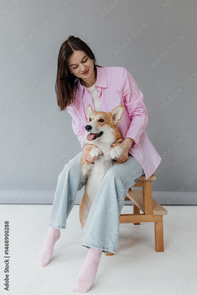 Happy brunette sitting next to corgi dog indoors. Cheerful girl in good mood stroking animal. Concept of friendship between man and pet.