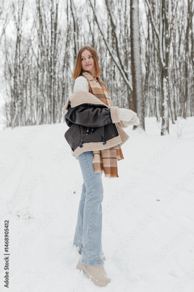 Happy smiling young woman wearing coat, scarf, and mittens enjoying winter weather while walking in snowy winter park. Stylish winter women's clothing.