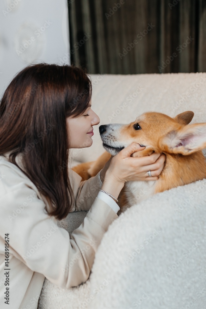 Happy brunette sitting next to corgi dog indoors. Cheerful girl in good mood stroking animal. Concept of friendship between man and pet.