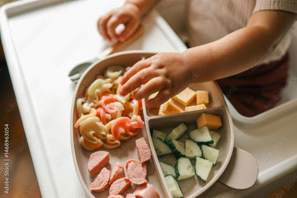 A toddler’s chubby hands grasp colorful pasta and cheese cubes from a divided plate during mealtime A toddler’s chubby hands grasp colorful pasta and cheese cubes from a divided plate during mealtime